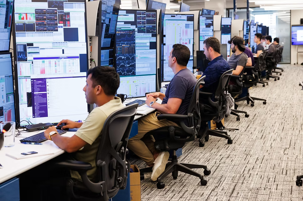 A long row of traders at curved multi-monitor stations on an open floor.